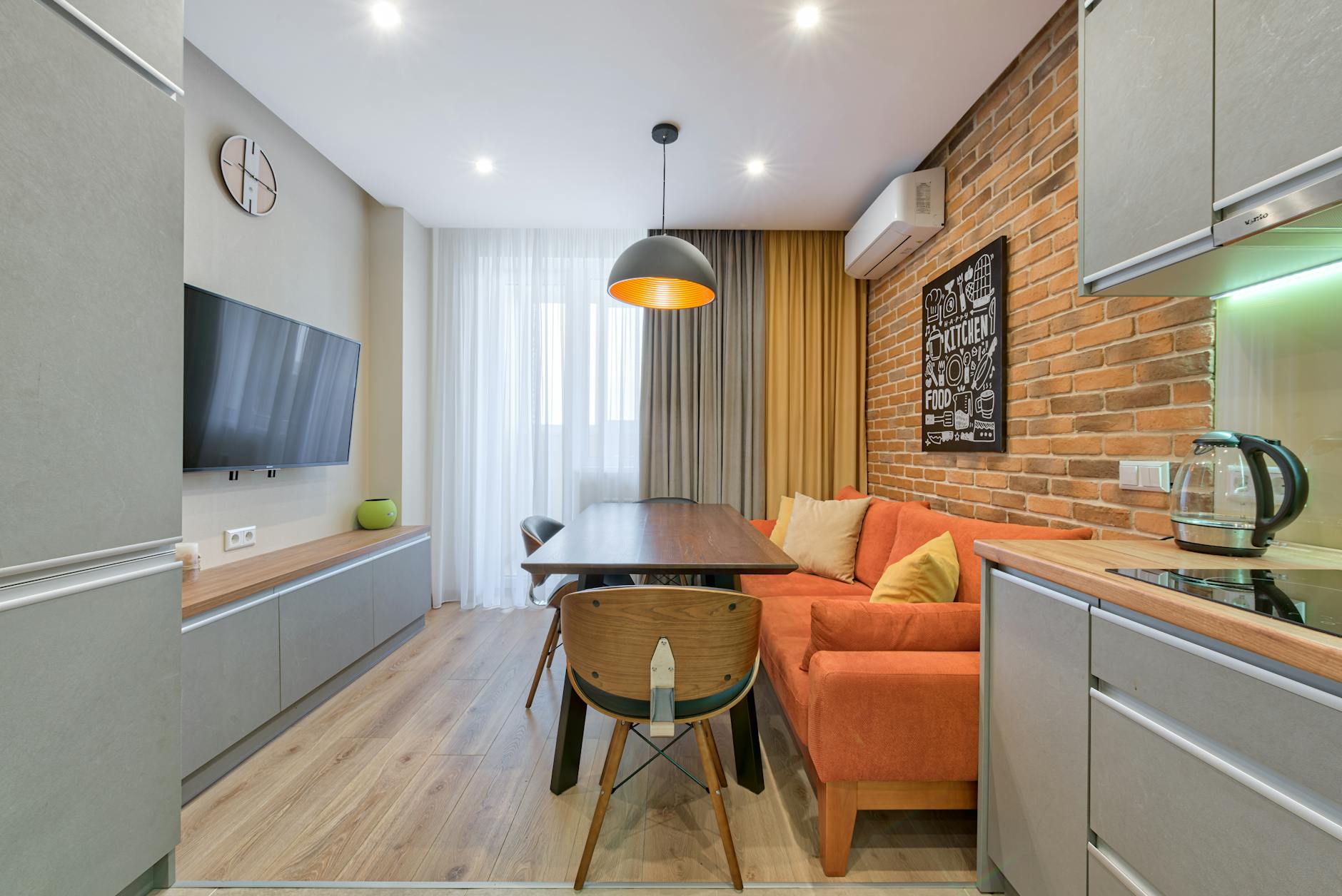 Small open-plan room with a dining table, orange corner sofa, brick wall and compact kitchen units