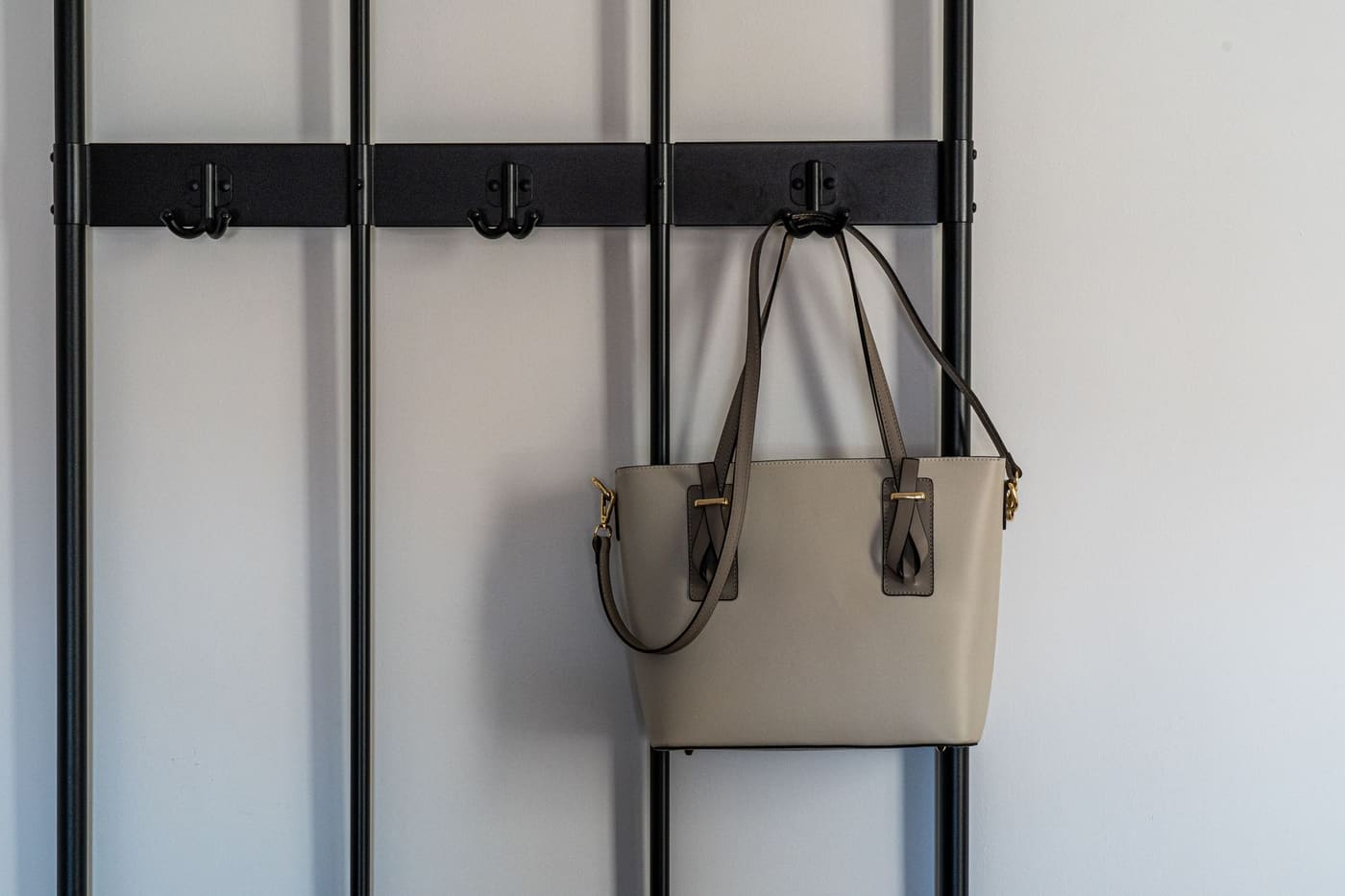 A beige handbag hanging from a black wall-mounted hook rail against a plain white wall