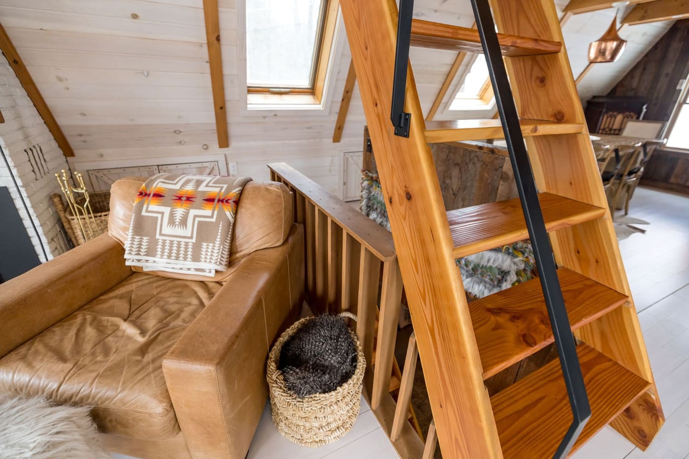 Wood-lined attic room with a steep wooden ladder up to a loft beside a leather armchair under roof windows