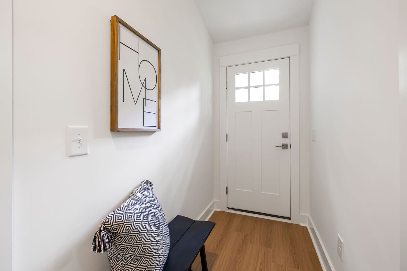 Narrow white entry hall with a front door, a framed print on the wall and a small bench with a cushion