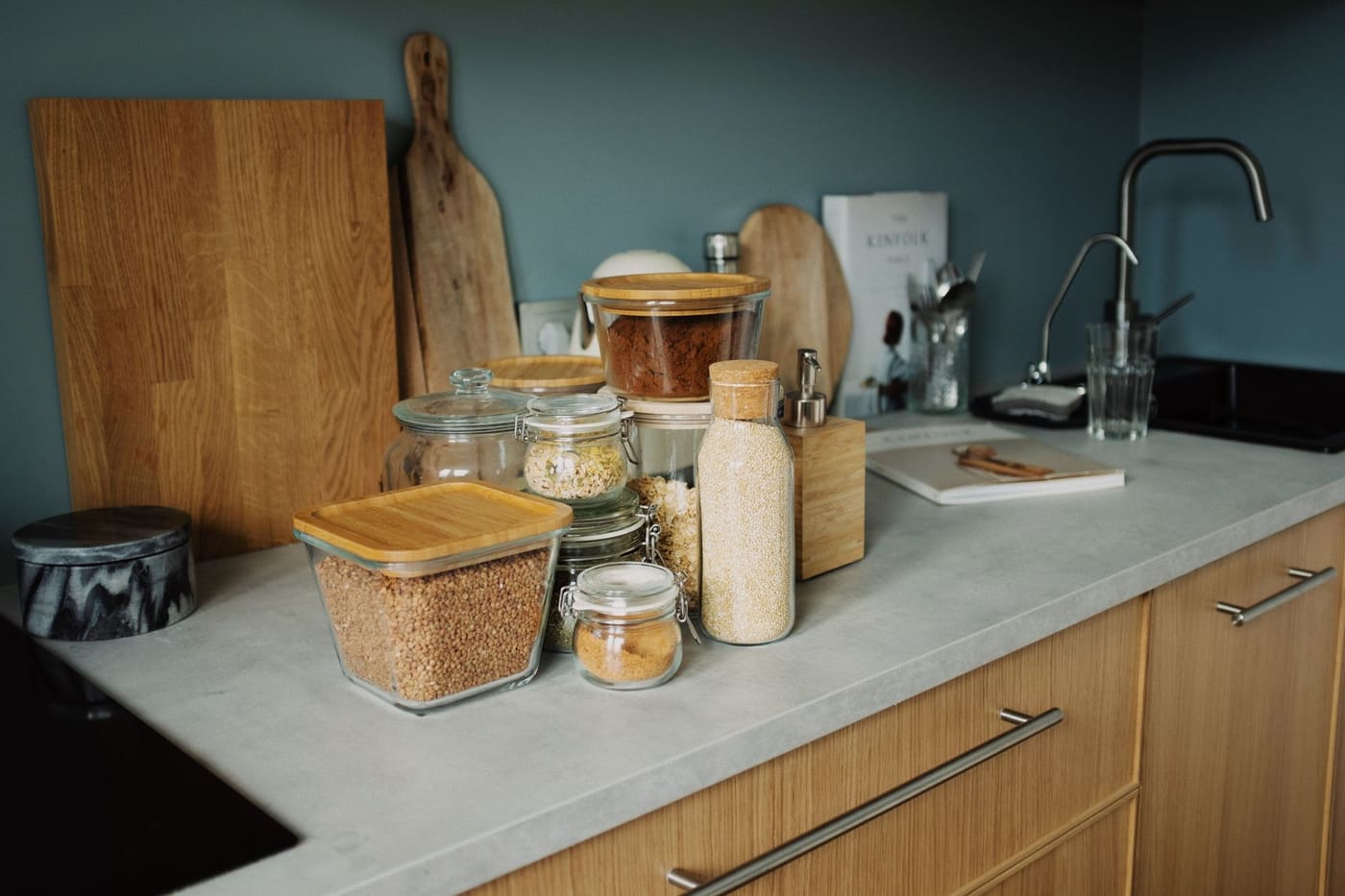 Kitchen counter with wooden cutting boards, glass storage jars and utensils beside a sink and under a wooden-front cabinet
