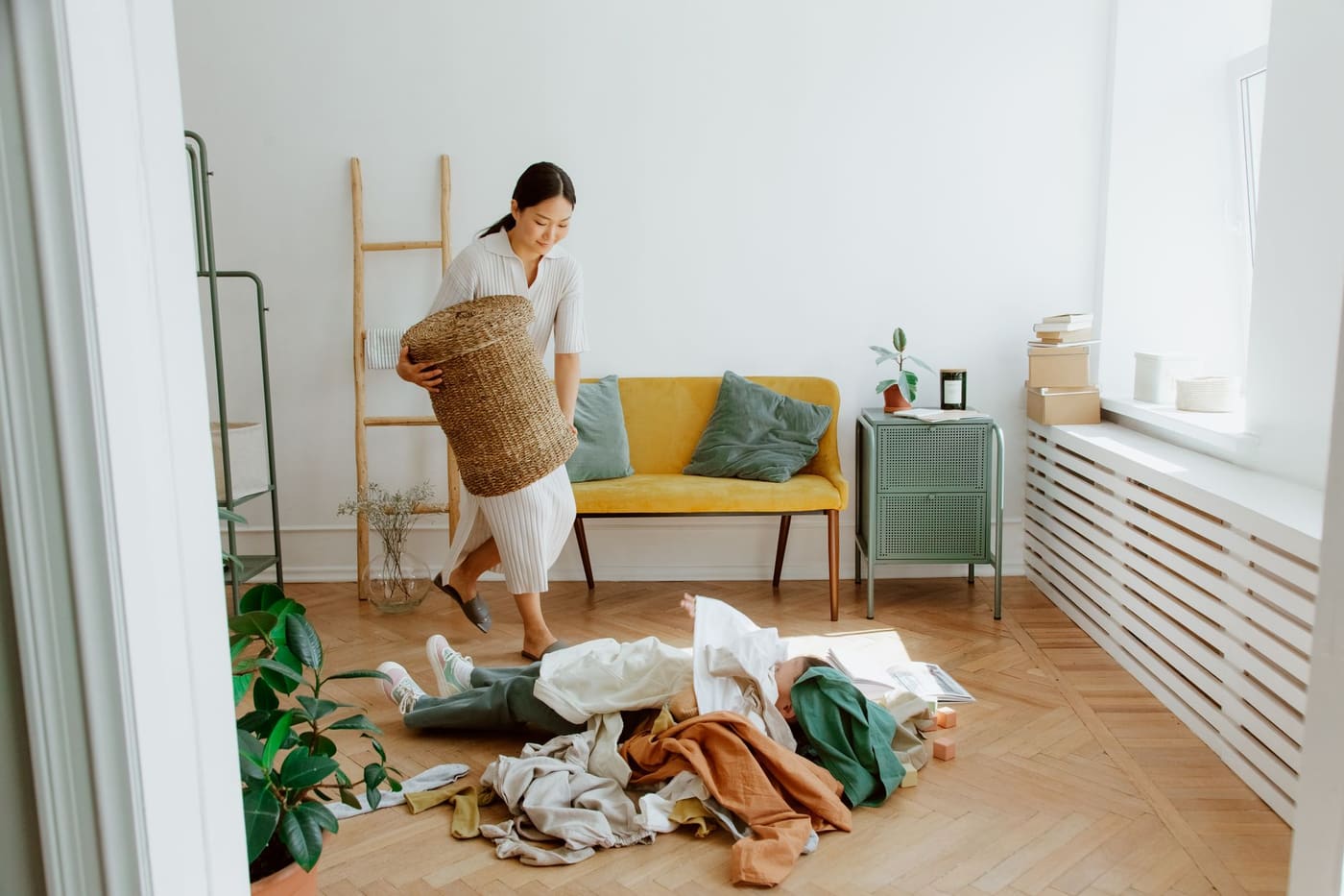 Bright living room with a woman carrying a basket past clothes piled on the wooden floor