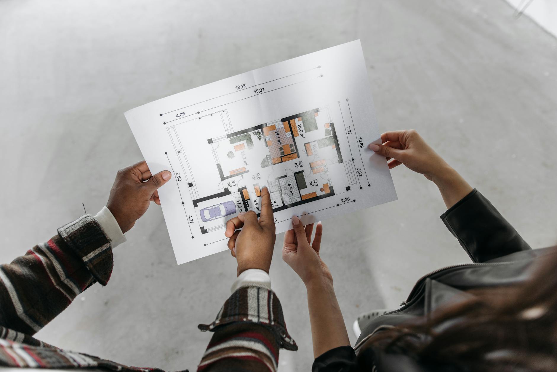 Two people holding a printed house floor plan, one pointing at a room